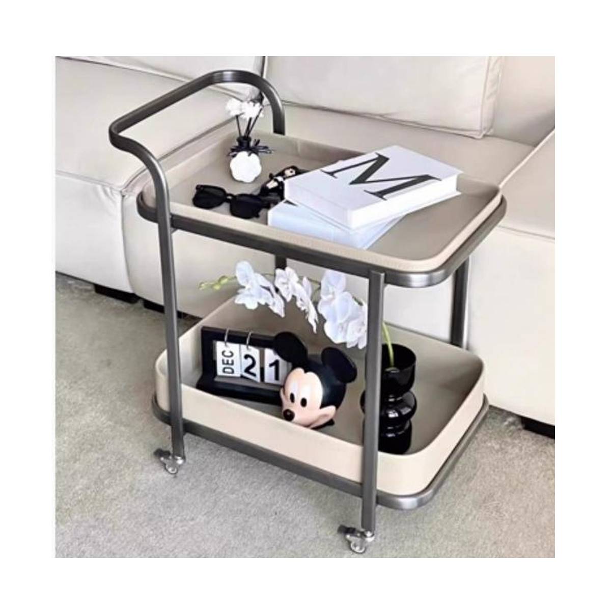 Beige rolling side table with stainless steel frame and saddle leather trays, styled with books, flowers, and a Mickey Mouse figurine.