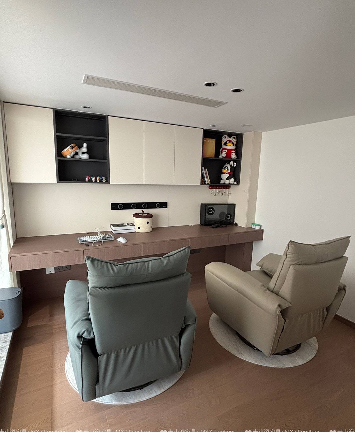 A home office setup featuring a long wooden desk, wall-mounted cabinets, and two swivel recliner chairs.