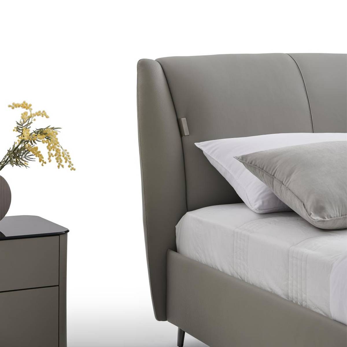 Side view of a grey upholstered bed with a curved headboard, white bedding, and a grey pillow, placed next to a bedside table with yellow flowers.