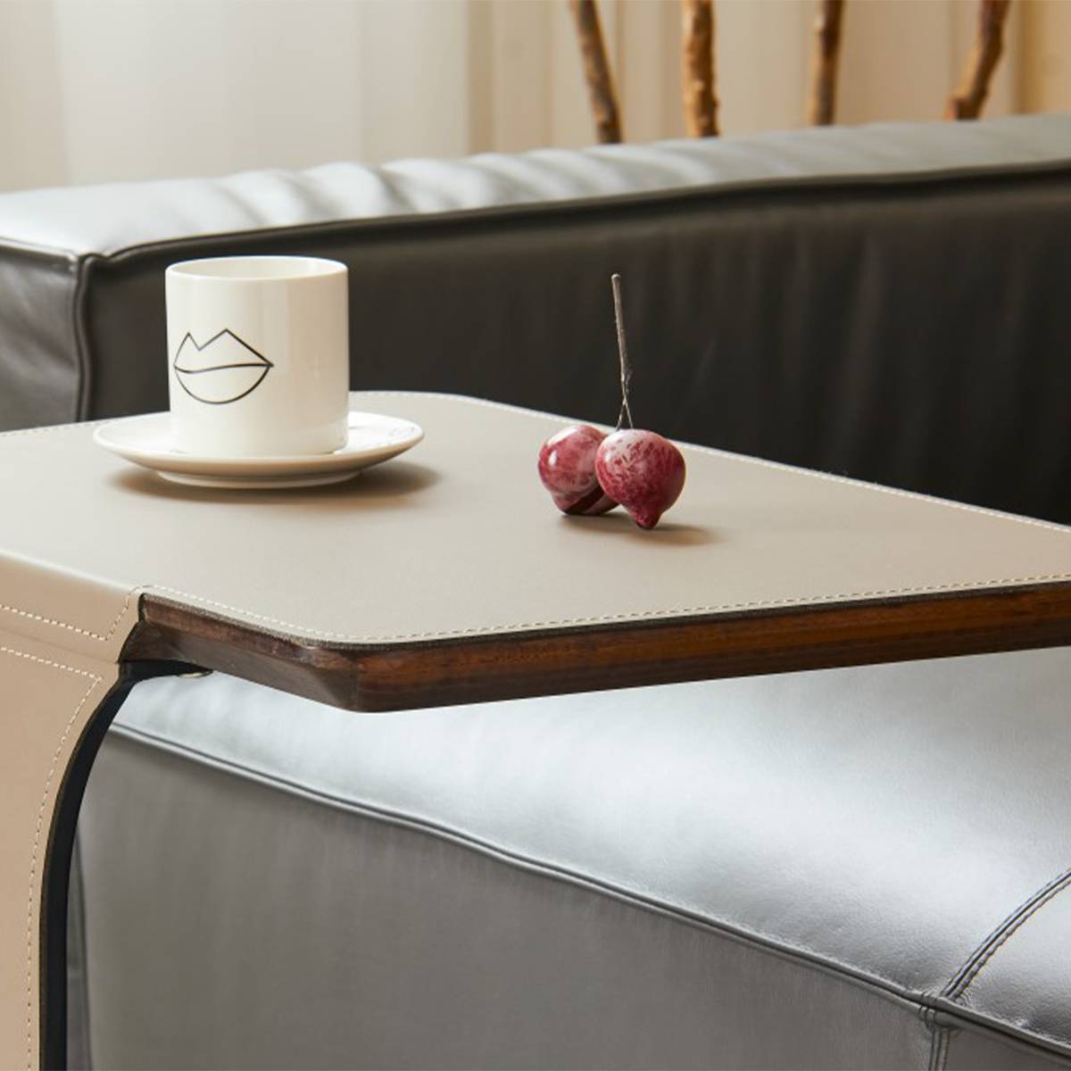 Close-up of a beige saddle leather side table top with a wooden edge, holding a white cup and cherries, placed next to a grey sofa.