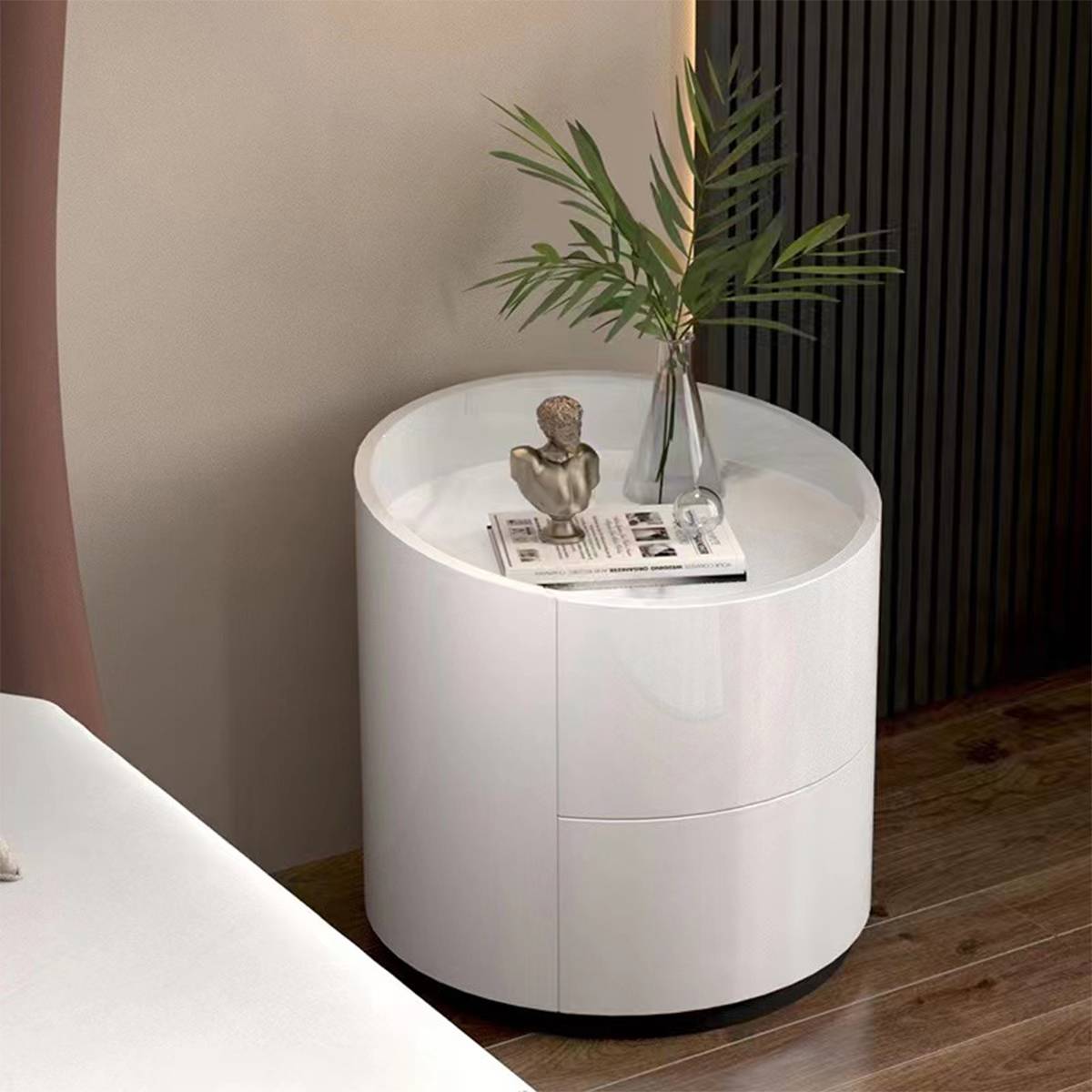 Cream white round bedside table with a drawer, featuring a plant and decorative bust, placed next to a bed.