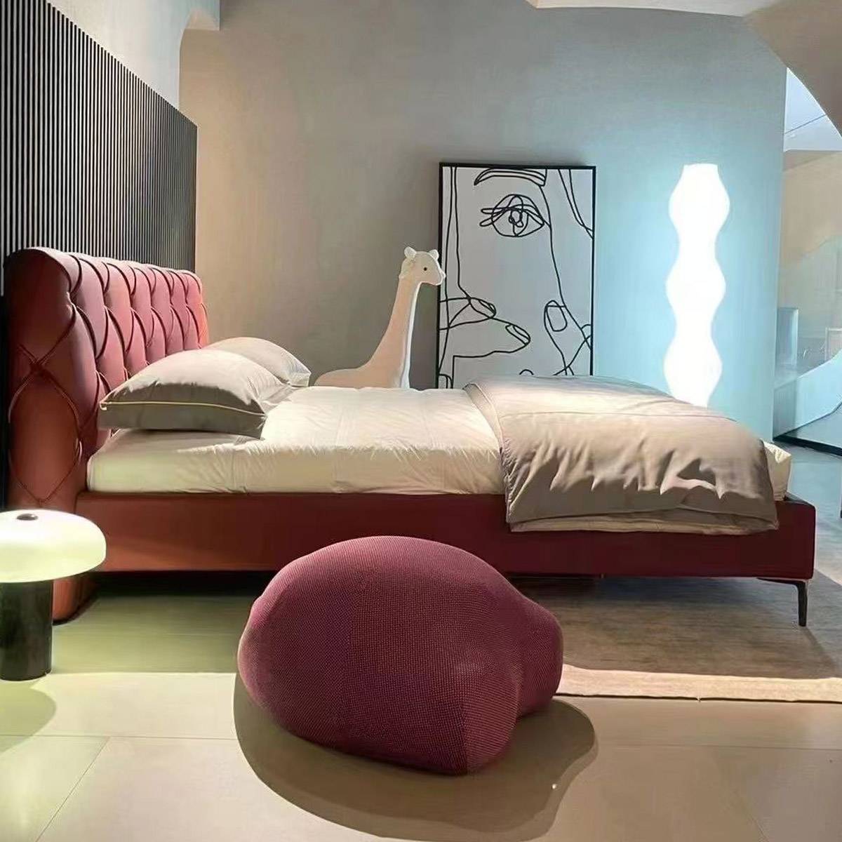 A burgundy tufted leather bed with a solid wood frame and metal legs, styled in a modern bedroom with a pink pouf and abstract art.