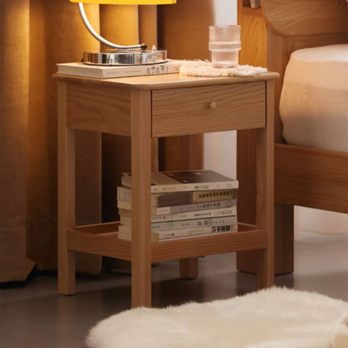 Light wood bedside table placed next to a bed with books on the lower shelf.
