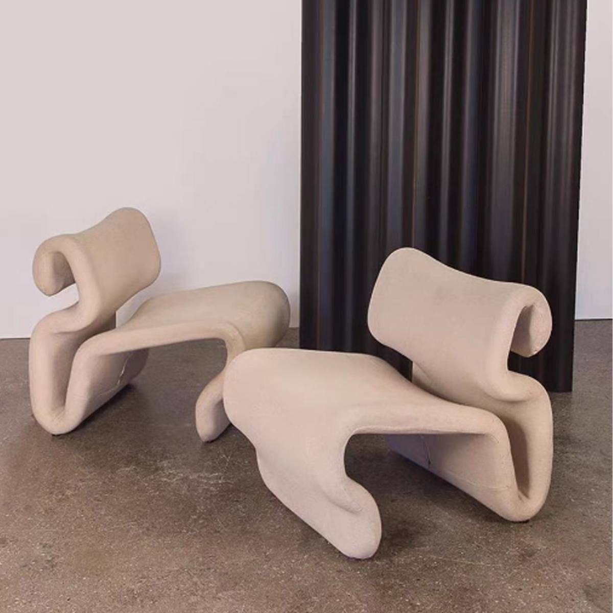 Two beige curved lounge chairs placed in a room with a concrete floor and dark curtain.