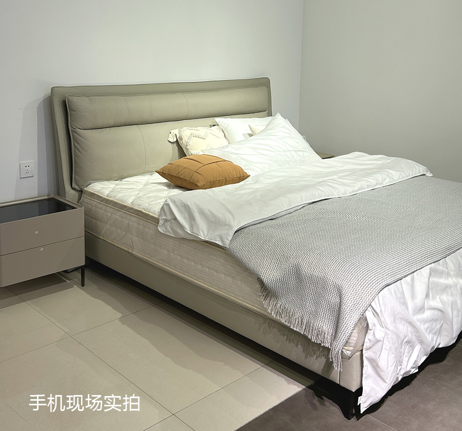 Real-life photo of the bed in a bedroom setting with bedding and a bedside table.